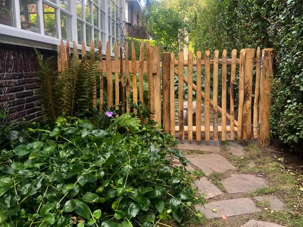 Franse rondhoutpoort van 80 cm bij 80 cm als doorgang naar een tuin met een hondje dat achter de poort zit.