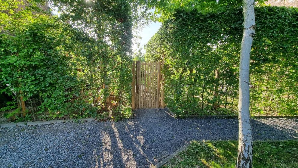 Franse rondhoutpoort van 150 cm hoog bij 80 cm breed als doorgang tussen een heg in de tuin.