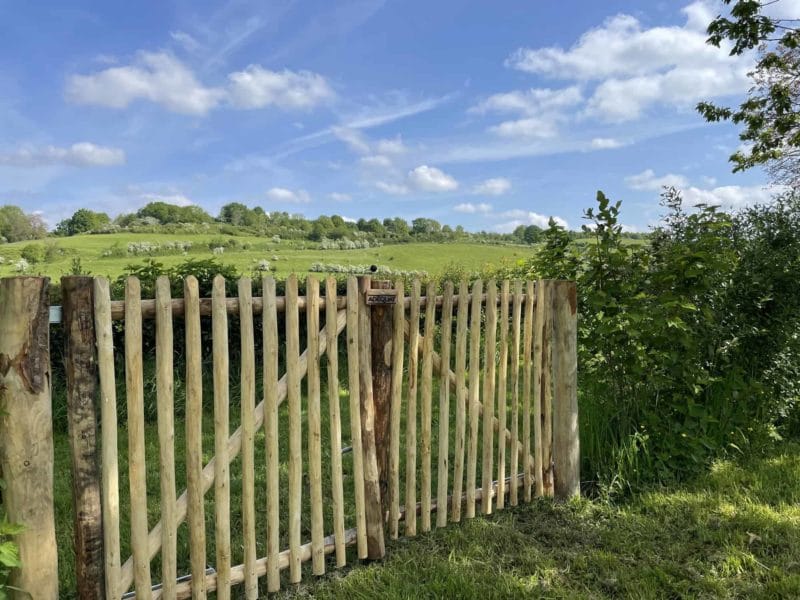 Dubbele franse rondhoutpoort van 100 cm breed en 100 cm hoog met een simpele sluiting aan de buitenkant. De foto is genomen vanaf de voorkant van de poort en je kijkt uit over een mooi groen landschap. De poort zorgt voor een nog landelijkere uitstraling.