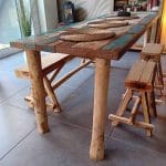 Boomstamkankje van kastanjehout van 150 cm breed en twee kastanjehouten krukjes. Deze staan bij een tafel binnen in de woning. De bankjes en de tafel stralen een Ibiza look uit.