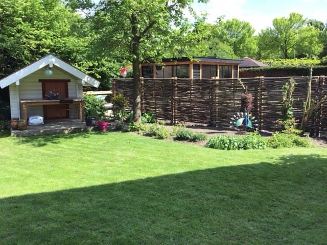 Hazelaarschermen somme horizontaal gevlochten gebruikt als creatieve en sfeervolle afscheiding tussen twee tuinen. De hazelaarschermen staan in de buurt van een tuinhuisje in een tuin met veel groen en gras.