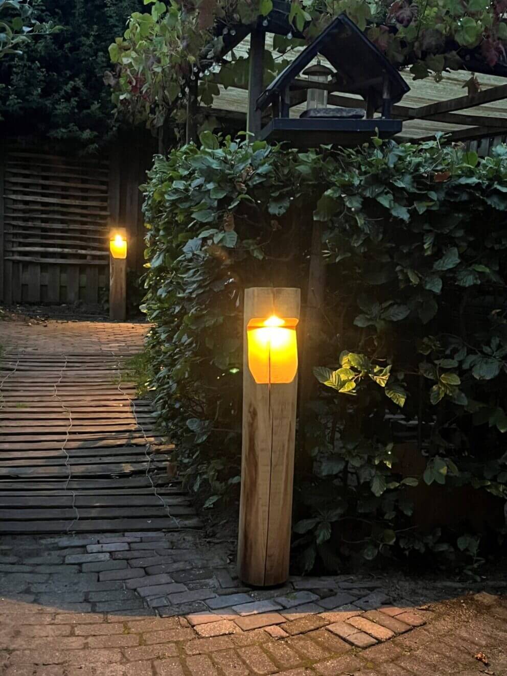 Kastanje looppad met houten op maat gemaakte tuinverlichting Bella in rustiek vierkant formaat, op voeten geplaatst (niet in de grond) met stopcontact outlet aan achterzijde. Kastanje hekwerk horizontaal bevestigd aan schutting.